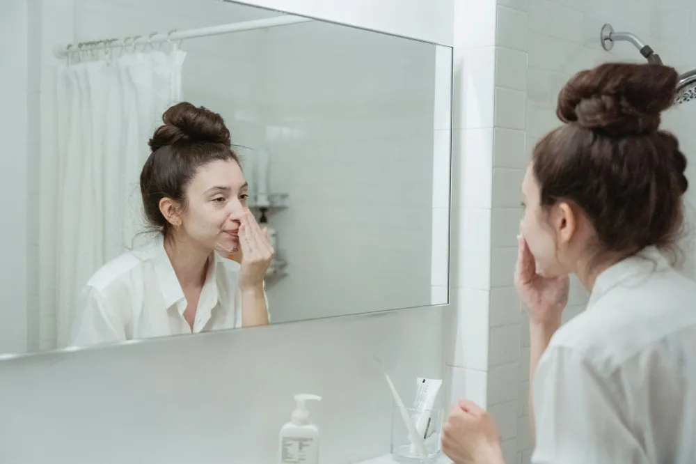 Woman examining under-eye area in bathroom mirror while holding skincare serum