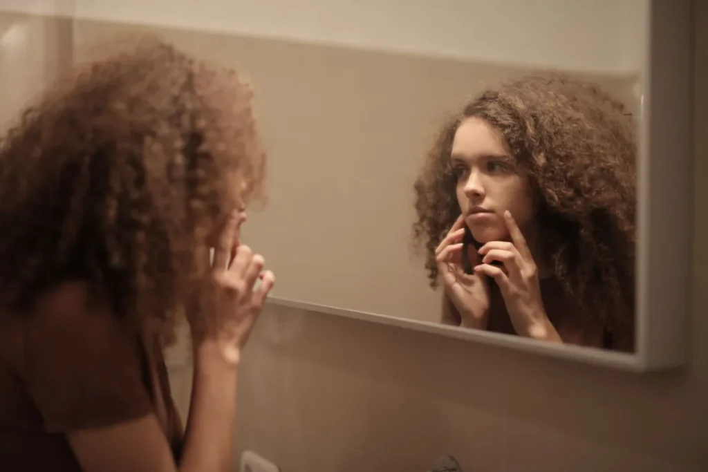 Woman closely examining red irritated skin in bathroom mirror after over-exfoliating